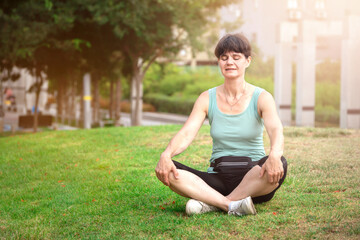 Middle aged smiling caucasian woman meditating or doing yoga on the grass in public park outdoors in summer sunny morning