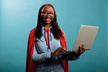 Positive and happy superhero woman wearing mighty hero red cape surfing internet on modern laptop...