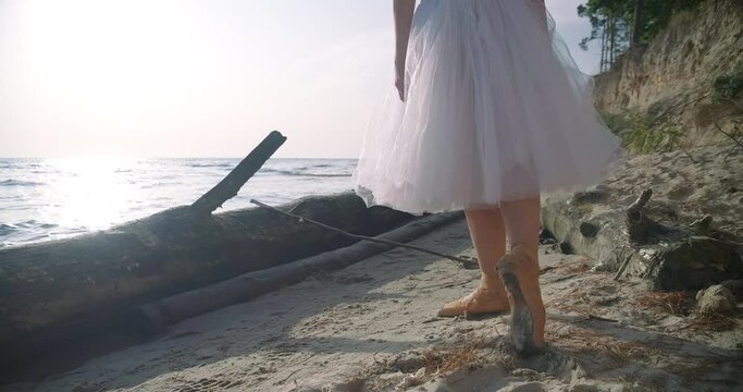 Tracking shot of slim graceful woman in pointies walking on sandy beach at dawn. Caucasian talented elegant ballerina in tutu strolling in slow motion on river bank at sunrise. Cinema 4k ProRes HQ