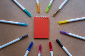 Red notepad for notes with red sheets. Blank space for notes. Mocap. On warm ochre kraft background. Small spring-loaded notebook resting on table surrounded by markers. Children's creativity, school