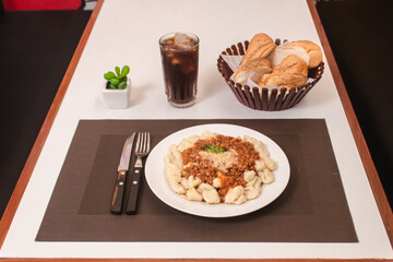 gnocchi with meat sauce with bread and soda on a white table.