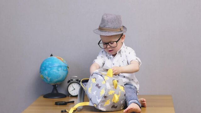 schoolboy concept smart baby boy child sitting in hat, glasses, packing schoolbag. elementary kindergarten kid starting school year, preparing first days at school. 
