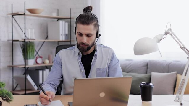 Handsome bearded man studying online using laptop web camera, writing lecture in notebook, talking with teacher using headset. Remote working,home education, job interview. Telework concept.