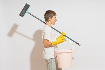 A teenager in rubber gloves with a floor brush on his shoulder and a bucket in his hands. Passes by a white wall. Cleaner concept.