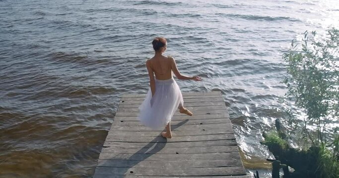 Wide shot gorgeous ballerina doing developpe and arabesque in slow motion in sunshine on wooden pier. Beautiful graceful Caucasian woman dancing on river bank in sunrays. Cinema 4k ProRes HQ