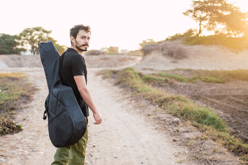 Young musician walking on a countryside road with a guitar on his shoulder. Looking to the camera