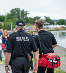 Two adult male medical first responders walking on a road among a crowd of people. Both are wearing black and grey colored paramedic uniforms with a soft red cloth medical first aid bag and radio.