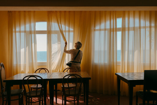 Man In Dining Room In Hotel With Warm Light Opening Curtains