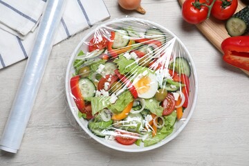 Bowl of fresh salad with plastic food wrap on white wooden table, flat lay - obrazy, fototapety, plakaty