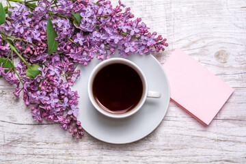 Obraz na płótnie Canvas White cup with coffee or tea, note with wishes, a bouquet of lilacs on a wooden background. Violet spring flowers. Morning composition.
