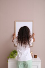 Woman hanging frame on pale rose wall over chest of drawers in room, back view