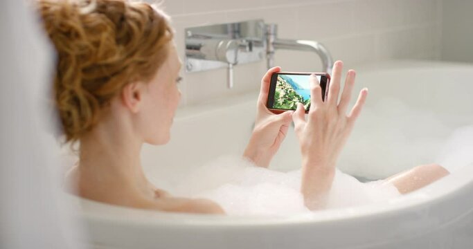 Relaxed woman in a bath with a phone looking at pictures of ex boyfriend while in hot water. Young female alone in a tub zoom in on digital photos. Pampering of a lady in a bathroom during a breakup