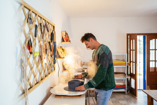 Happy Hispanic craftsman using hat steamer in workshop