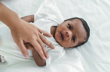 Fototapeta na wymiar Closeup portrait of cute African american newborn baby lying on bed and looking at camera in bedroom. Mother hand lulling little child to sleeping. Child Care, Black family, Newborn baby boy. New life