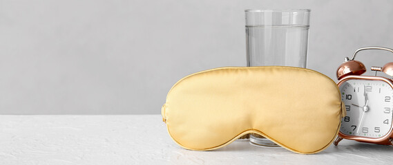 Fototapeta na wymiar Sleep mask, glass of water and alarm clock on light background with space for text