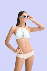 Fototapeta premium Young tanned woman in swimsuit and sunglasses on lilac background