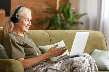 Mature female soldier in headphones using laptop at home