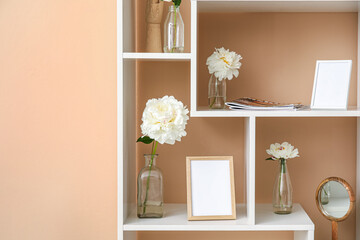 Shelf unit with blank photo frames and beautiful peony flowers near beige wall, closeup
