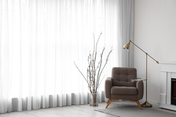 Fototapeta premium Vase with tree branches, armchair and lamp near light curtain in living room