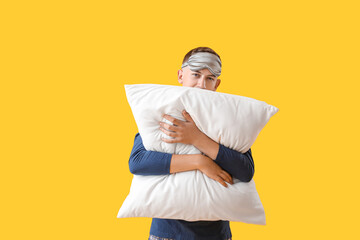 Young man with sleeping mask hugging pillow on yellow background