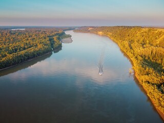 River valley with a steep bank with a forest of trees, sunlight and a boat on the water