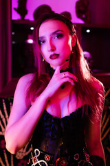 Portrait of a beautiful girl in a corset. Purple lighting. She looks seductively into the camera, holding her hand near her face. Halloween concept, nightclub, masquerade