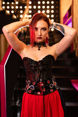 Beautiful girl in a corset stands on the background of the stairs. Holds her hair in her hands, looks intimidatingly into the camera. Halloween concept, nightclub, masquerade