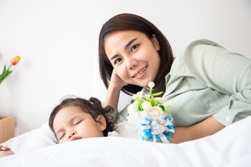 Obraz na płótnie Canvas Mother’s day. Loving mom carying of her daughter at home. Bright portrait of happy mum holding jasmine flowers near little girl sleeping on bed.