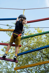 Child in safety harness and helmet in rope park. Boy climbs attached with carbine to cable on rope...