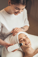 Beautician with ultrasonic scrubber cleansing woman's face skin at spa salon. Electrical equipment...