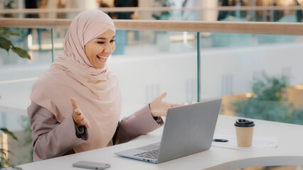 Fototapeta na wymiar Happy young arab businesswoman sitting at office desk typing on laptop working on internet checking new app smiling looking at camera showing thumb up approval sign consent support symbol high rating