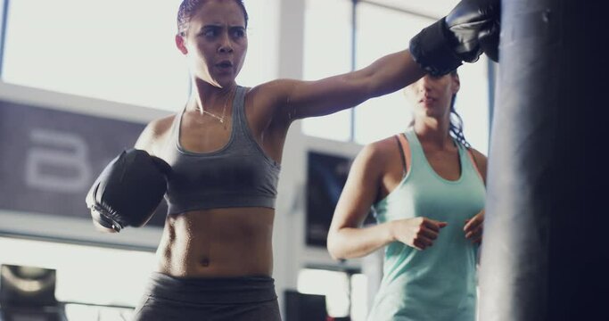 Female athlete boxer, exercising with a punching bag with coach watching at training fitness gym. Active sport woman with boxing gloves doing workout fighting routine with instructor indoors.