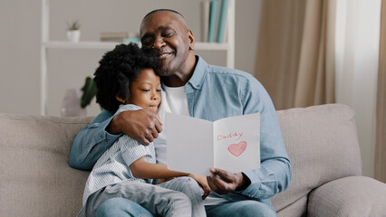 Mature african american man with cute kid girl hugging sitting in room on sofa father reads greeting card loving daughter made gift with own hands for birthday or father's day dad happy cuddling baby