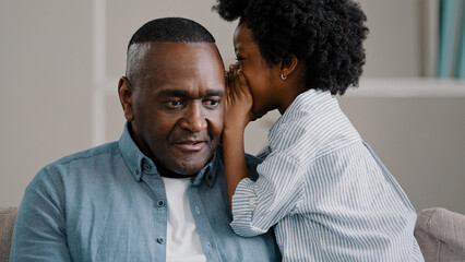 Little African American girl whispers in ear reveals secret to daddy daughter shares secrecy gossip confidential information mature father sitting on couch surprised from conversation opens eyes wide