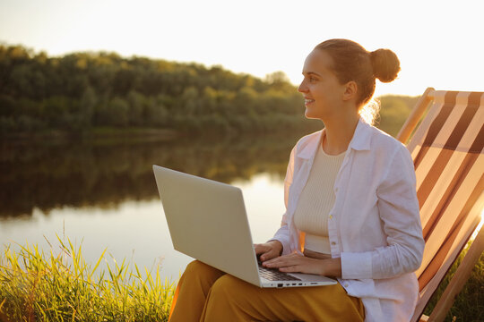 Side View Portrait Of Smiling Woman With Bun Hairstyle Wearing White Shirt Sitting By The Water In Folding Chair And Working On Laptop, Looking Away Enjoying Nature And View.