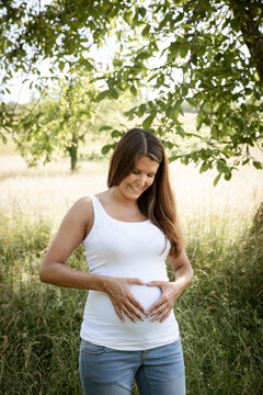 pretty attractive young pregnant woman with white top is standing in high meadow and forming a heart with her hands on her belly out in the nature