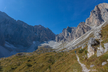 Fototapeta na wymiar Path at the foots of rocky ridges of Croda Rossa di Sesto and Cima Undici Mountains in Comelico region with lonely mountaineer in the middle of green meadows, Dolomites, Italy