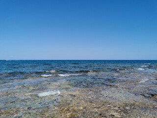 The coast of the Mediterranean Sea, waves, clear water through which the bottom is visible. All this against a blue sky with clouds.