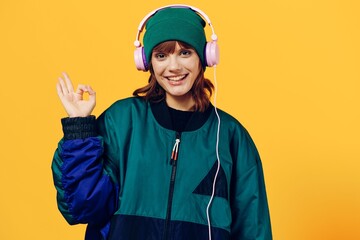 a joyful woman in a green jacket and a hat in the style of the 90s enjoys music standing in pink headphones on a yellow background showing the ok sign while looking at the camera