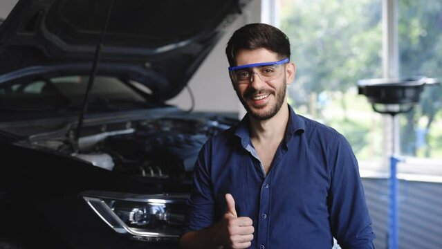 Portrait of young caucasian bearded handsome man auto mechanic or manager in goggles smiling to camera and giving thumb up in garage. Cheerful guy standing at cars service