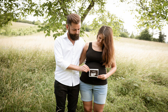 beautiful young pregnant woman with her husband standing in green meadow holding ultrasonic picture of her baby