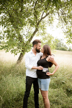 beautiful young pregnant woman with her husband standing in green meadow holding ultrasonic picture of her baby
