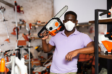 African-american man in face mask standing in salesroom of gardening tools shop with chainsaw in...
