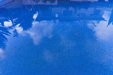 Swimming pool with blue water on a sunny day. Houses and palm trees are reflected in the water. The tile is visible through a transparent clean aod. July in summer. Spain.