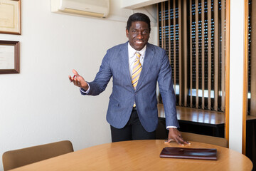 Friendly african american entrepreneur standing in meeting room near table with folder of documents, inviting to negotiations
