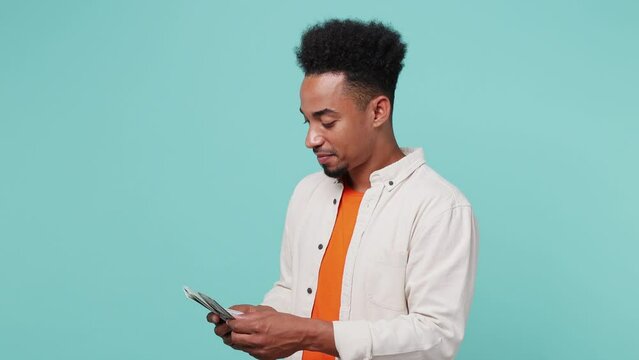 Young satisfied fun happy man of African American ethnicity 20s he wearing shirt t-shirt hold stack of cash money in dollar banknotes count salary isolated on plain pastel light blue cyan background