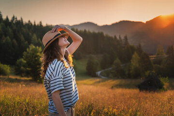 Young beautiful happy cheerful woman in a hat enjoying the sunset in a meadow with mountains view