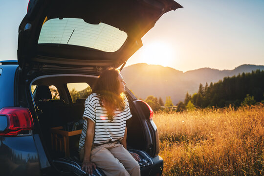 Young cheerful woman traveler sitting in open trunk of car enjoying sunset in mountains
