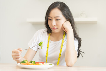 Obraz na płótnie Canvas Diet in bored. Closeup broccoli unhappy beautiful asian young woman, girl on dieting, hand holding fork in salad plate, dislike or tired with eat fresh vegetables. Nutrition of healthy food good taste