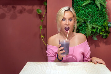 Young woman looking surprised at her smoothie while sitting in a coffee shop. Healthy lifestyle concept.